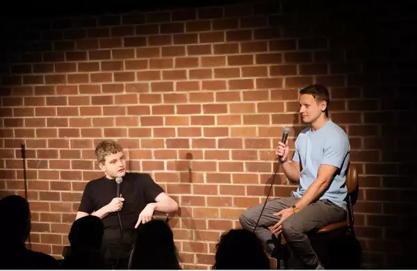 Wheels and the Legman performing on stage holding microphones. Behind them is a brick wall.