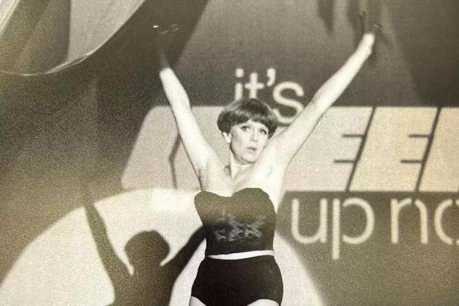 Black-and-white photo of Lois Weaver in a strapless bodysuit with arms raised high on stage, in front of a backdrop with partially visible words 'It's Queer Up North'