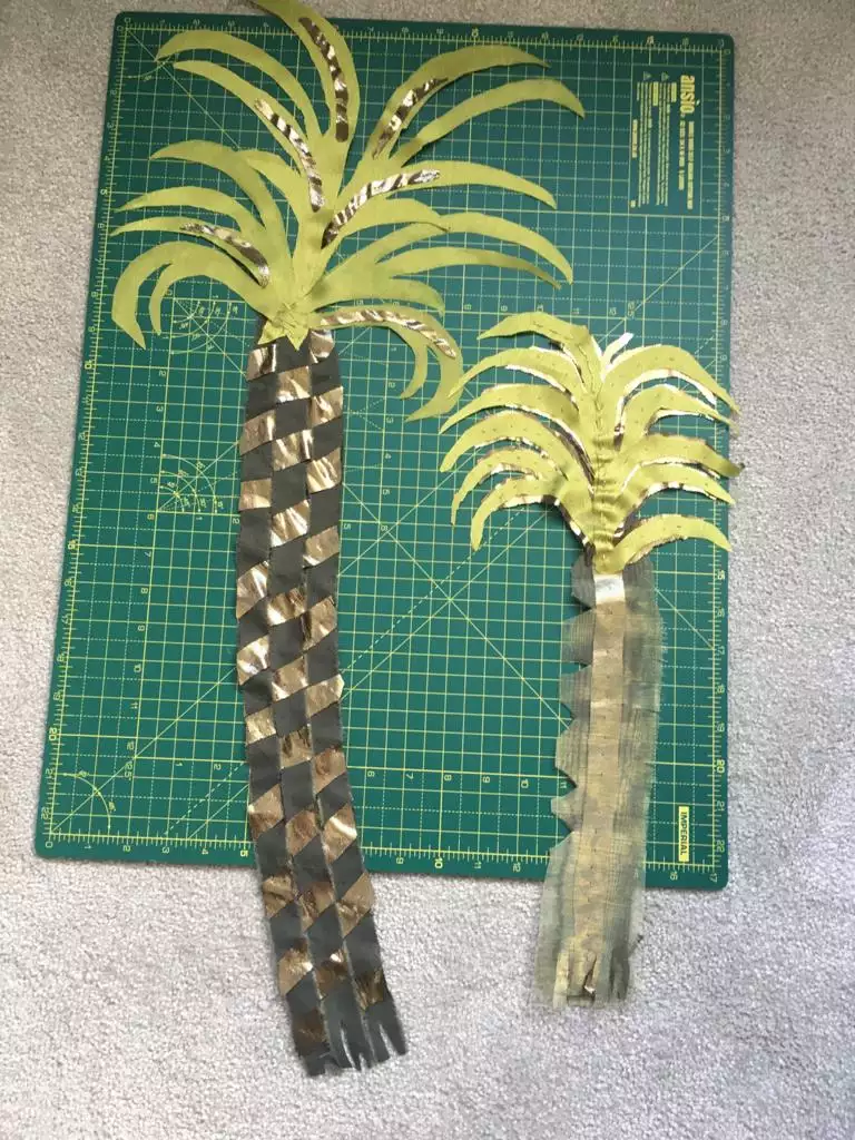 Two palm trees made from woven fabric strips in green, brown and copper laid out on a cutting mat.
