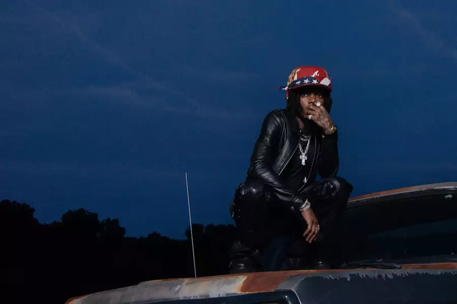 J.I.D wearing a black leather outfit and red hat. He squats on top of an old car at dusk.