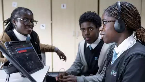 Three students in school uniforms sit around a tablet on a desk; one wears headphones and speaks, while the other two listen attentively. They are in a classroom with light wood cabinets in the background.