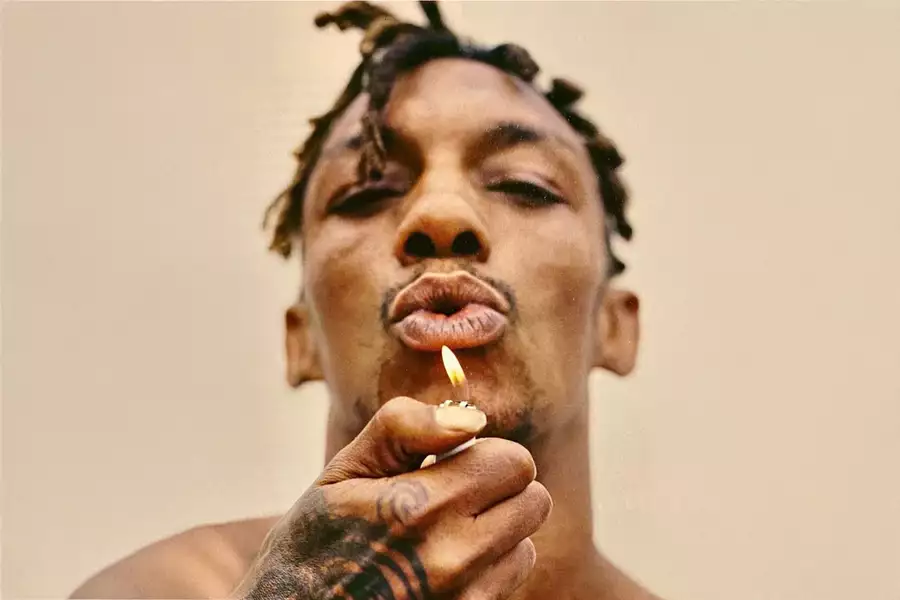 Tricky holds a lighter flame near his mouth, on a beige background.
