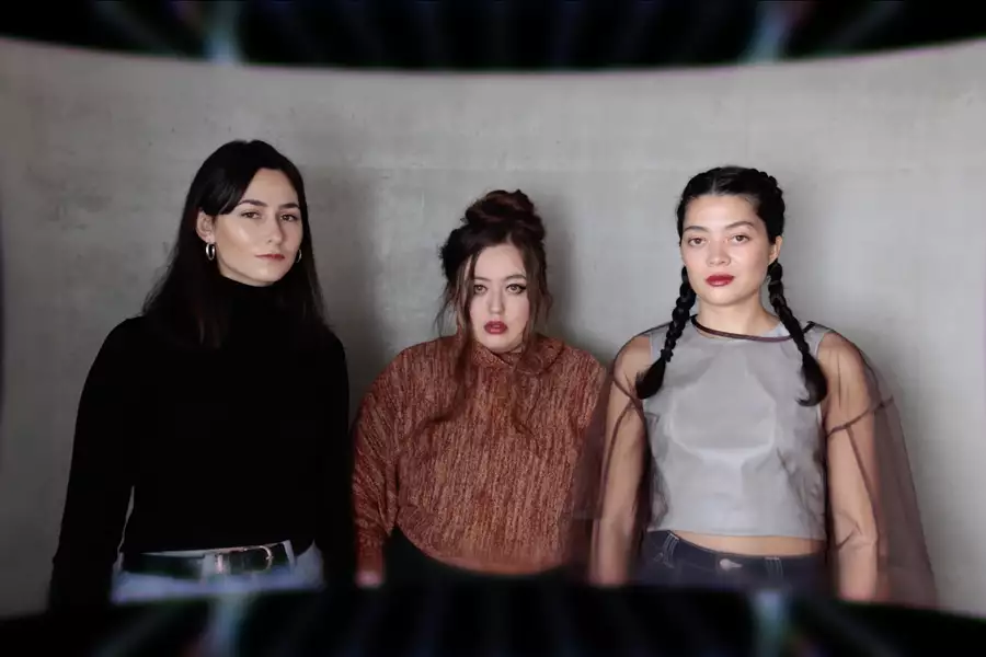 Three members of Keiken staring at the camera, stood in front of a large, grey piece of material.