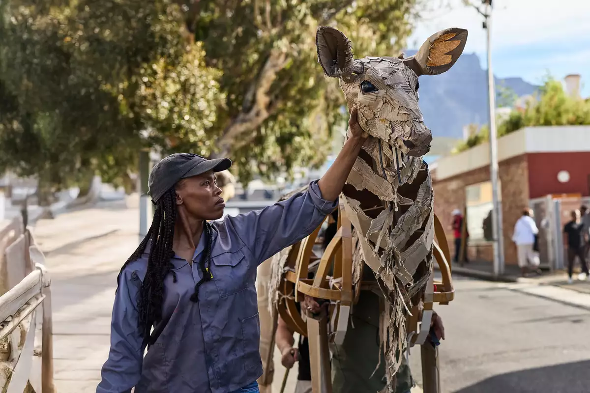 A woman in a cap touches the head of a life-sized giraffe puppet made from cardboard and wood, as part of The Herds. The background shows an urban environment with people watching and a scenic view of trees and mountains.