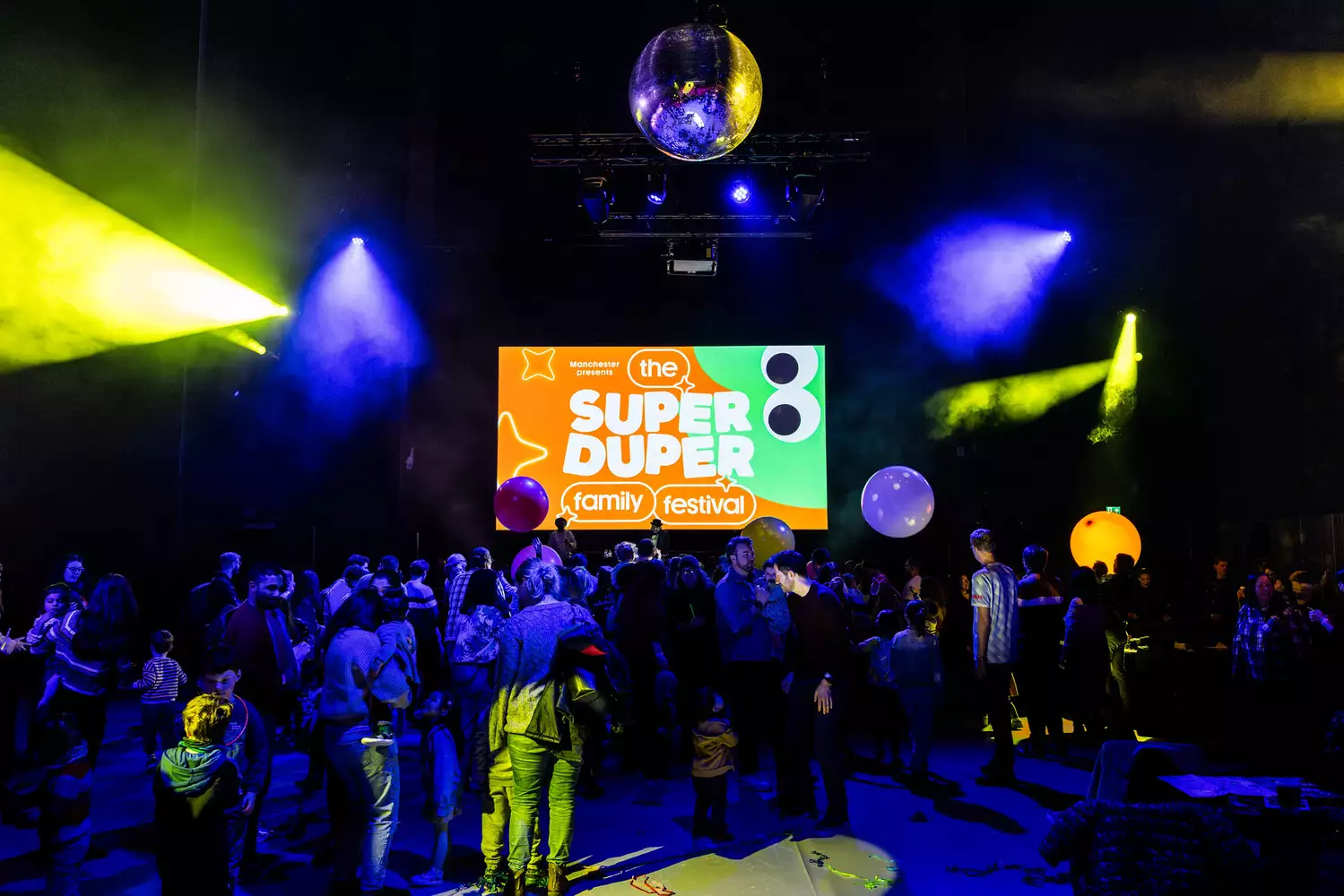A large crowd of families dancing in the Warehouse with a screen that reads 'Super Duper Family Festival'