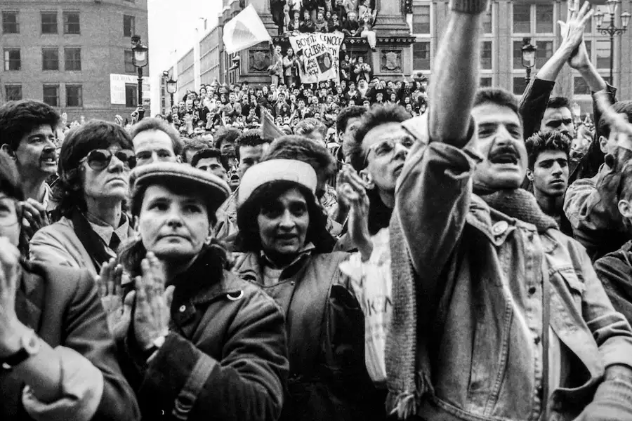 A large crowd of people, bundled in coats, clap, cheer, and hold signs at a protest. The photo is black and white.