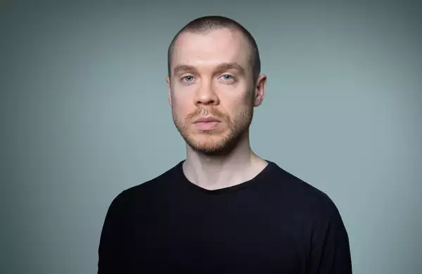 A headshot of Gary Stevenson with short hair and a neatly trimmed beard, wearing a plain black T-shirt. The background is a smooth gradient of gray-green.