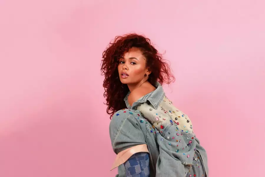 Savanna Darnell poses against a pink background, wearing an oversized embellished denim jacket and matching jeans. She crouches stylishly, showcasing blue patterned boots while gazing confidently at the camera.
