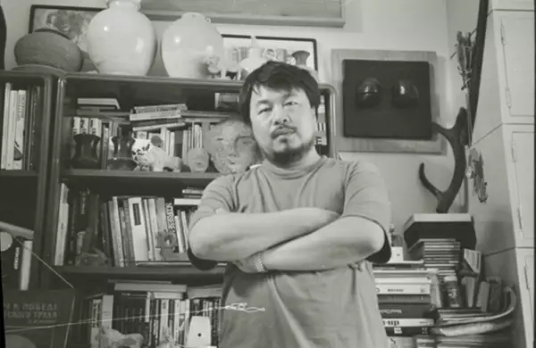 Black and white photograph of Ai Weiwei stood in front of a bookshelf