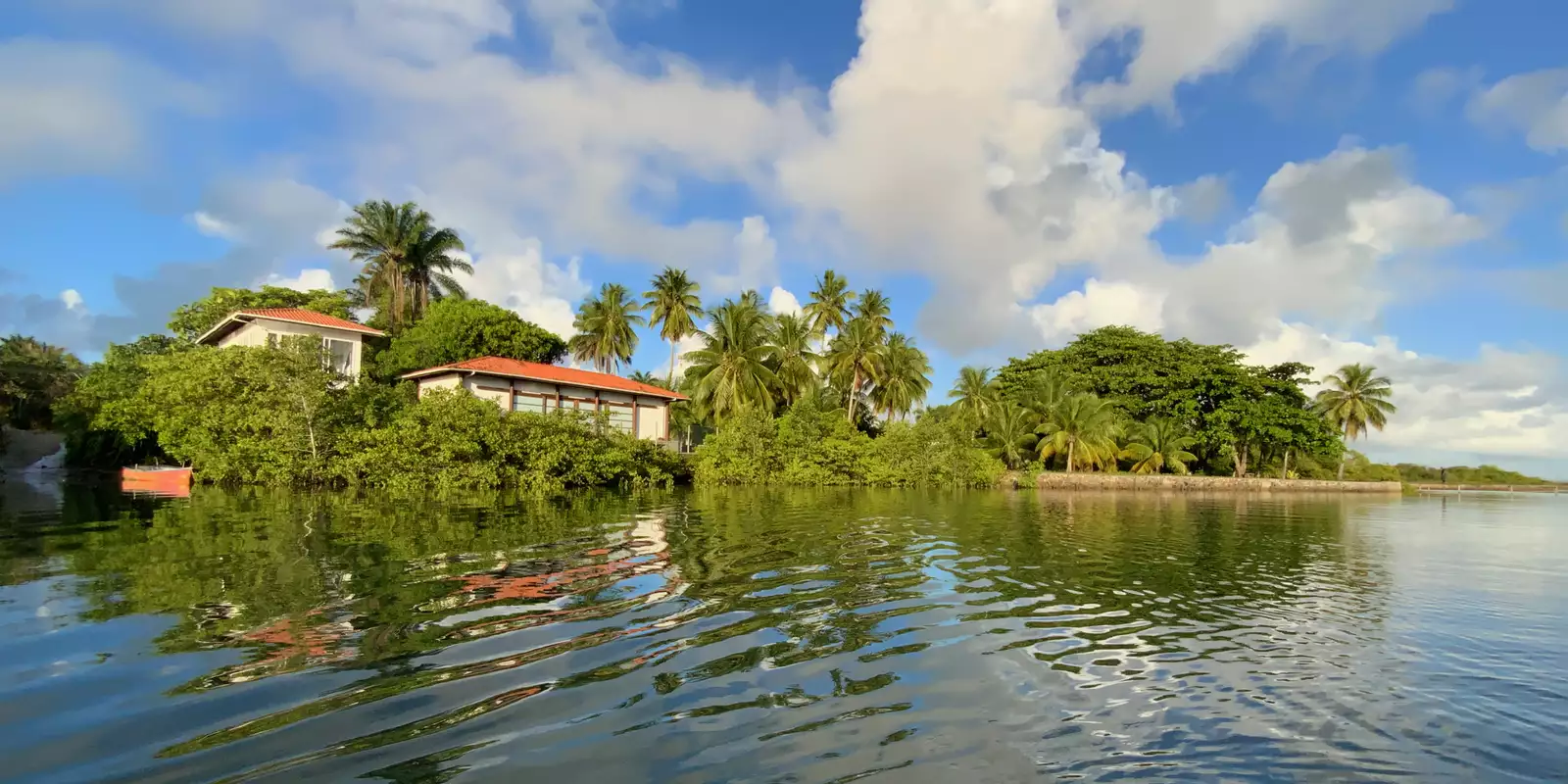 A tranquil waterfront scene shows Sacatar Institute nestled among palm trees and greenery, with reflections shimmering on the calm water. The sky is partly cloudy and sunny.