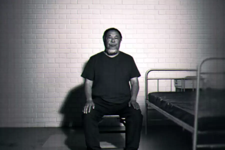 Black-and-white image of Ai Weiwei sitting on a chair beside a simple metal bed against a brick wall.