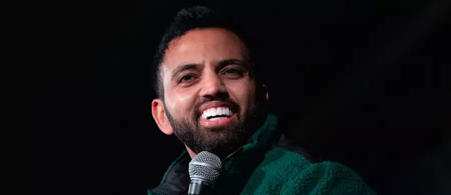Akaash Singh speaking into a microphone on a black background, wearing a green fleece