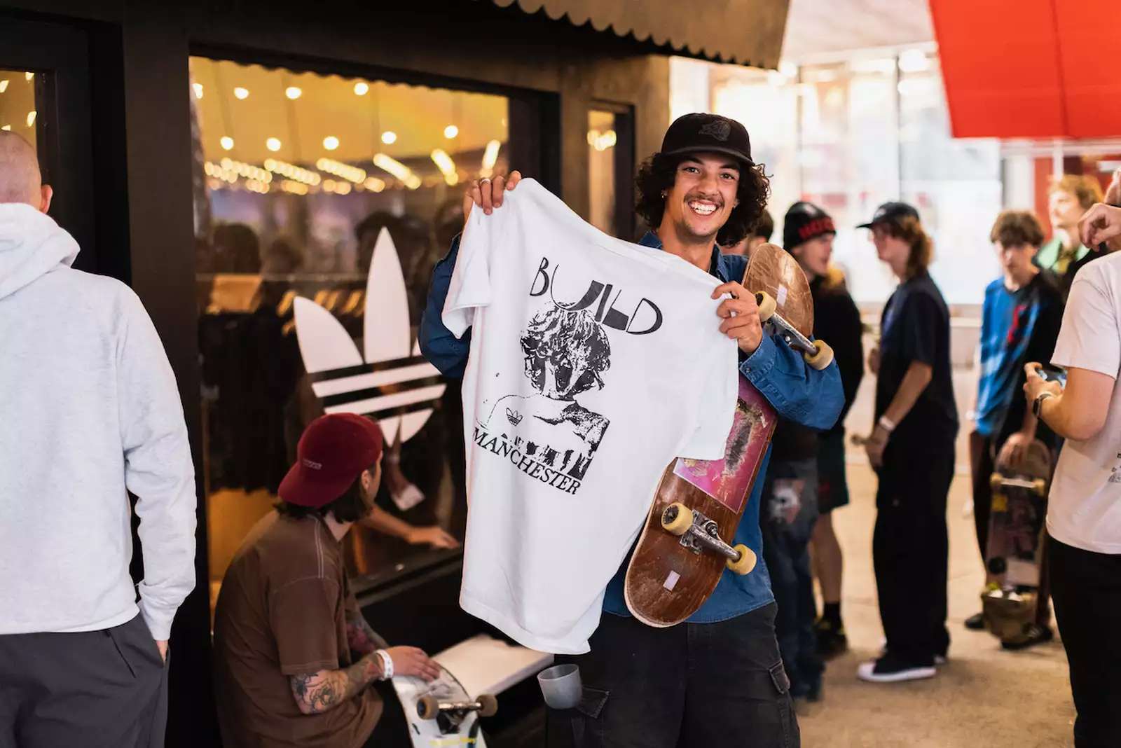 A person smiling and holding a skateboard and white t-shirt which says 'Build Manchester' on