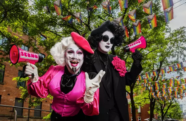Anna Phylactic and Liquorice Black pose on Manchester's Canal Street. There are colourful pride flags above them. They are holding pink branded microphones.