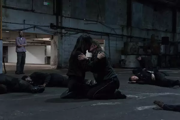 A photo of two people kneeling in large warehouse space embracing while other people lay on the floor around them