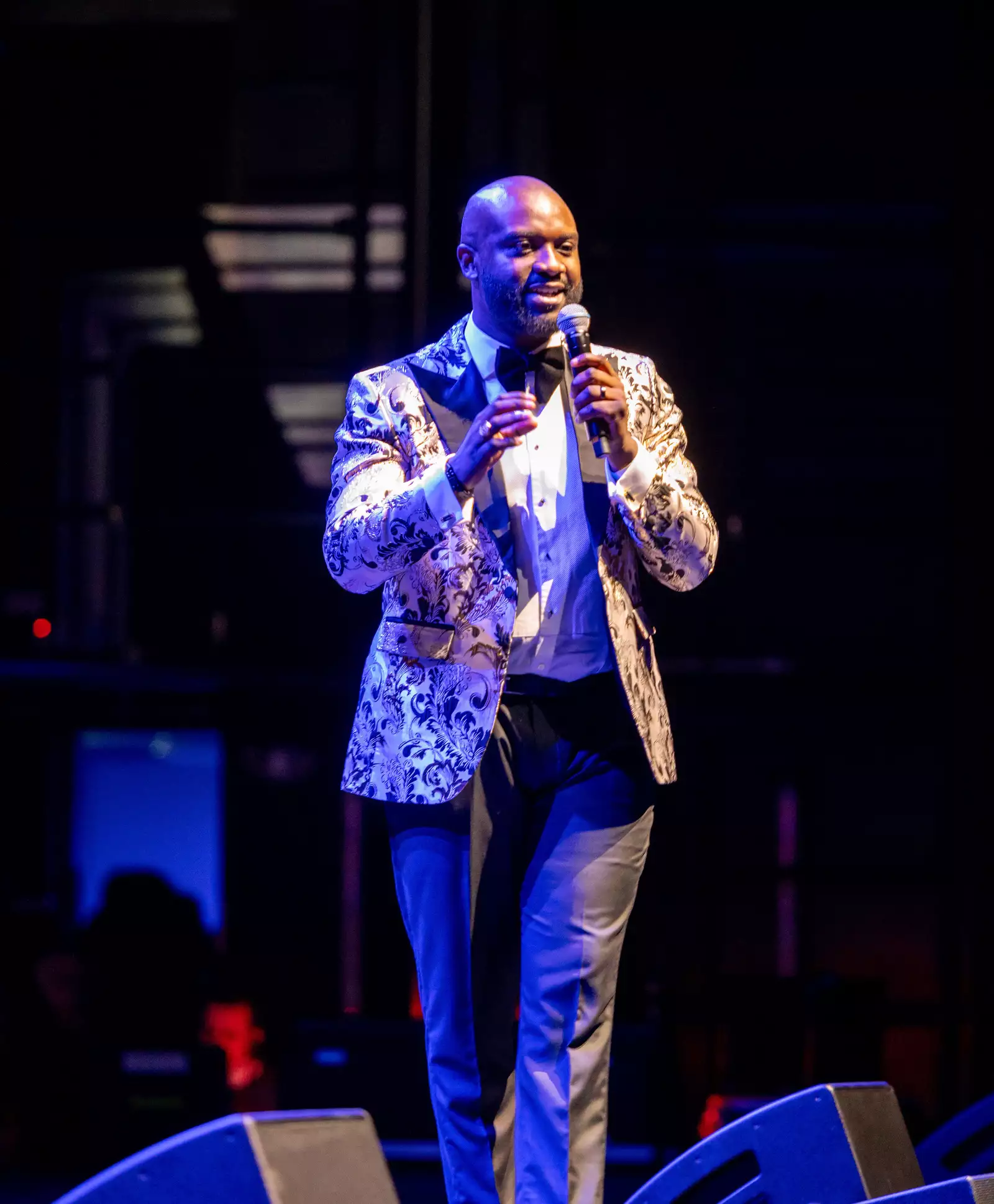 A man speaks into a microphone on stage at Black Creative Trailblazers. He is wearing a patterned silver suit and the lighting is blue.