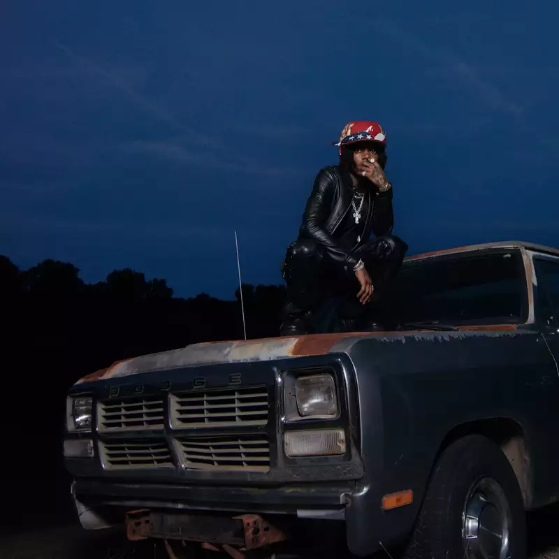 J.I.D wearing a black leather outfit and red hat. He squats on top of an old car at dusk.