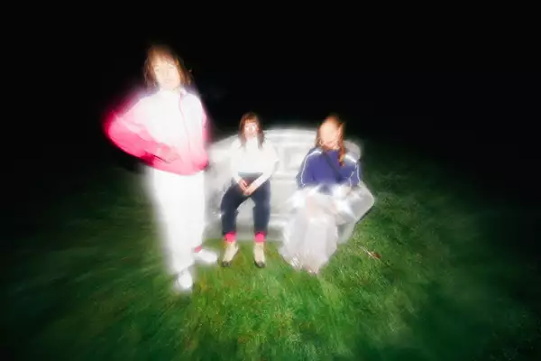 Blurry photo of three members of Stealing Sheep sat on a sofa outside in a grassy field