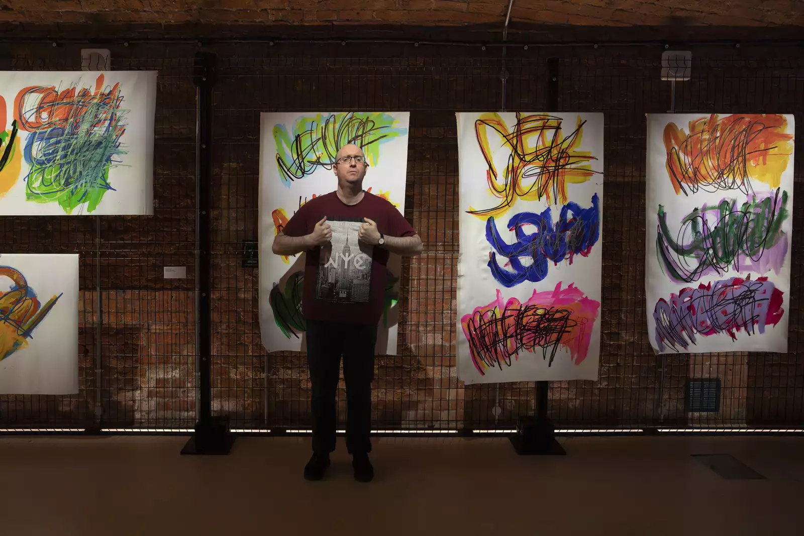 A photo of artist Michael Beard standing in front of a gallery wall of his paintings which feature brightly coloured layers of paint used to write the names of cities in a repetitive pattern