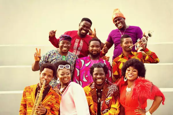 Nine members of Adédèji smiling with their instruments, wearing orange, purple and yellow clothes