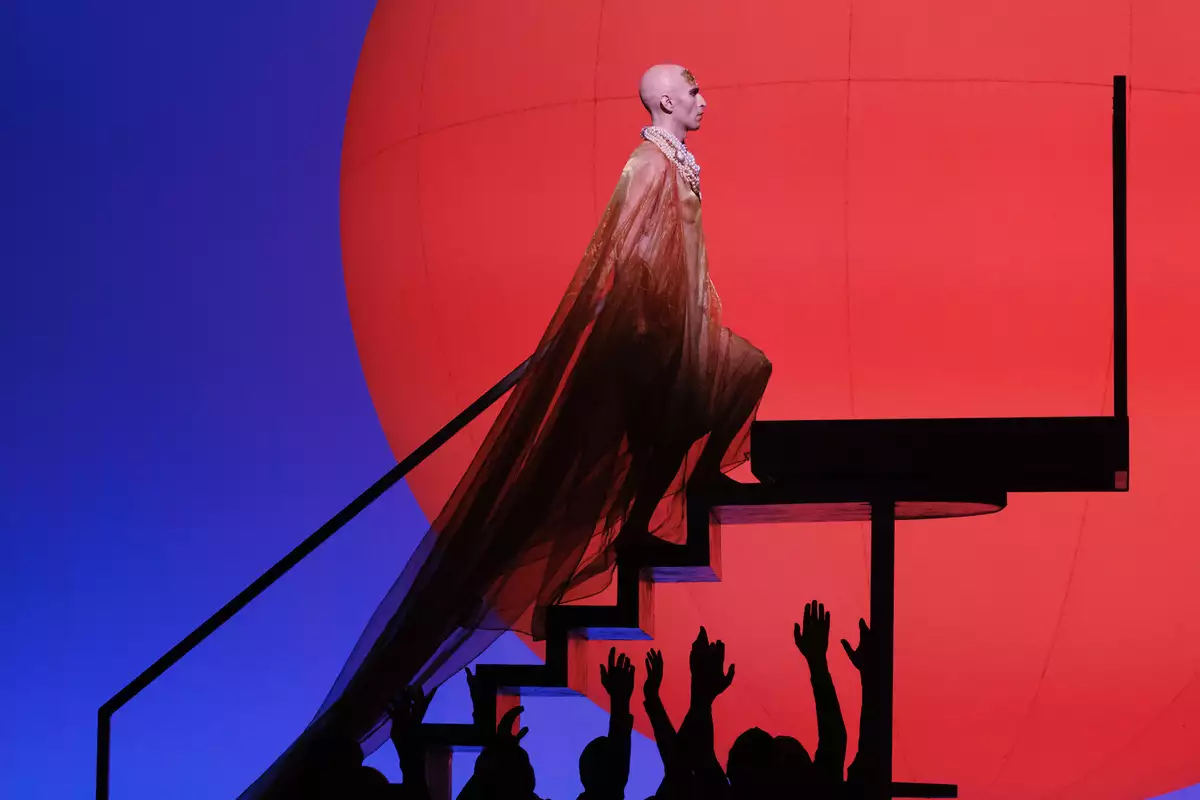 An opera stage, showing one person climbing steps in an opulent dress with others raising their hands towards them. The background is red and blue.