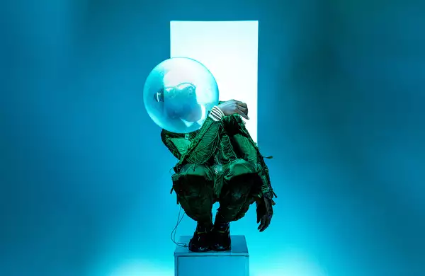 Benji Reid crouched on a small pedastal in front of a white strip against a blue background. He is wearing a green pattered suit and a bubble sculpture on his head.