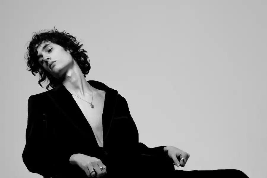 Black and white image of sombr wearing a black suit sitting on a chair