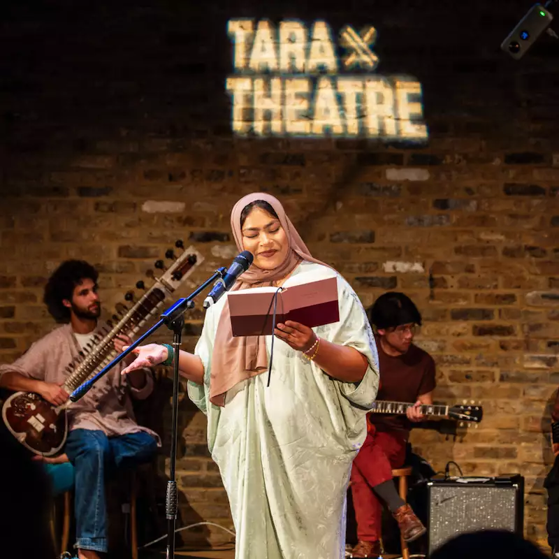 Nasima Bee on stage reading into a microphone. There are two musicians on stage and the words 'Tara x Theatre' are projected on the brick wall.