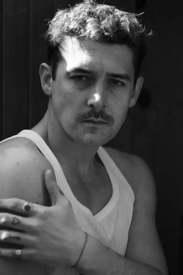 Black and white headshot of Greig Matthew wearing a white vest