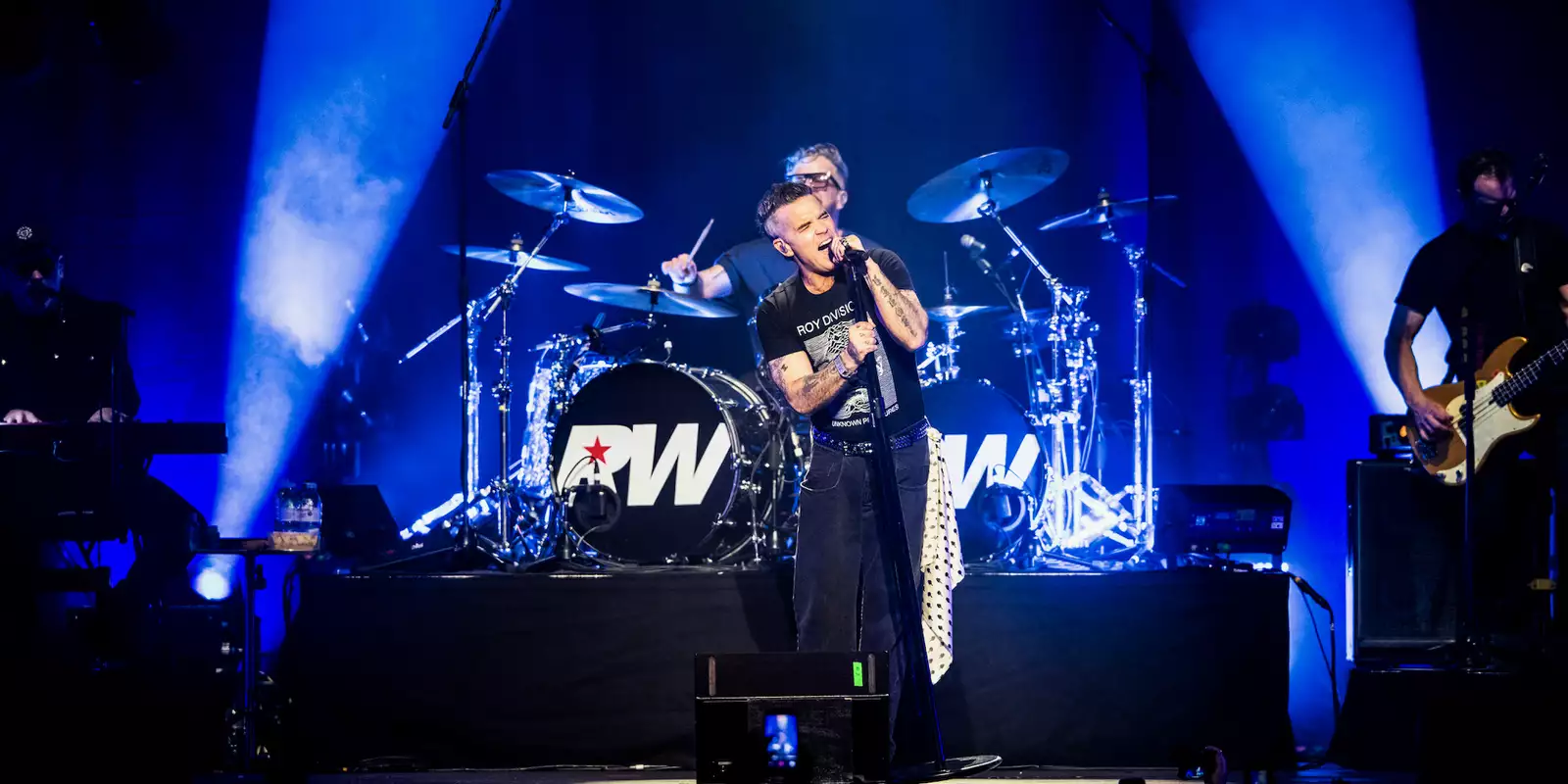 Robbie Williams on stage singing with blue lighting and his band
