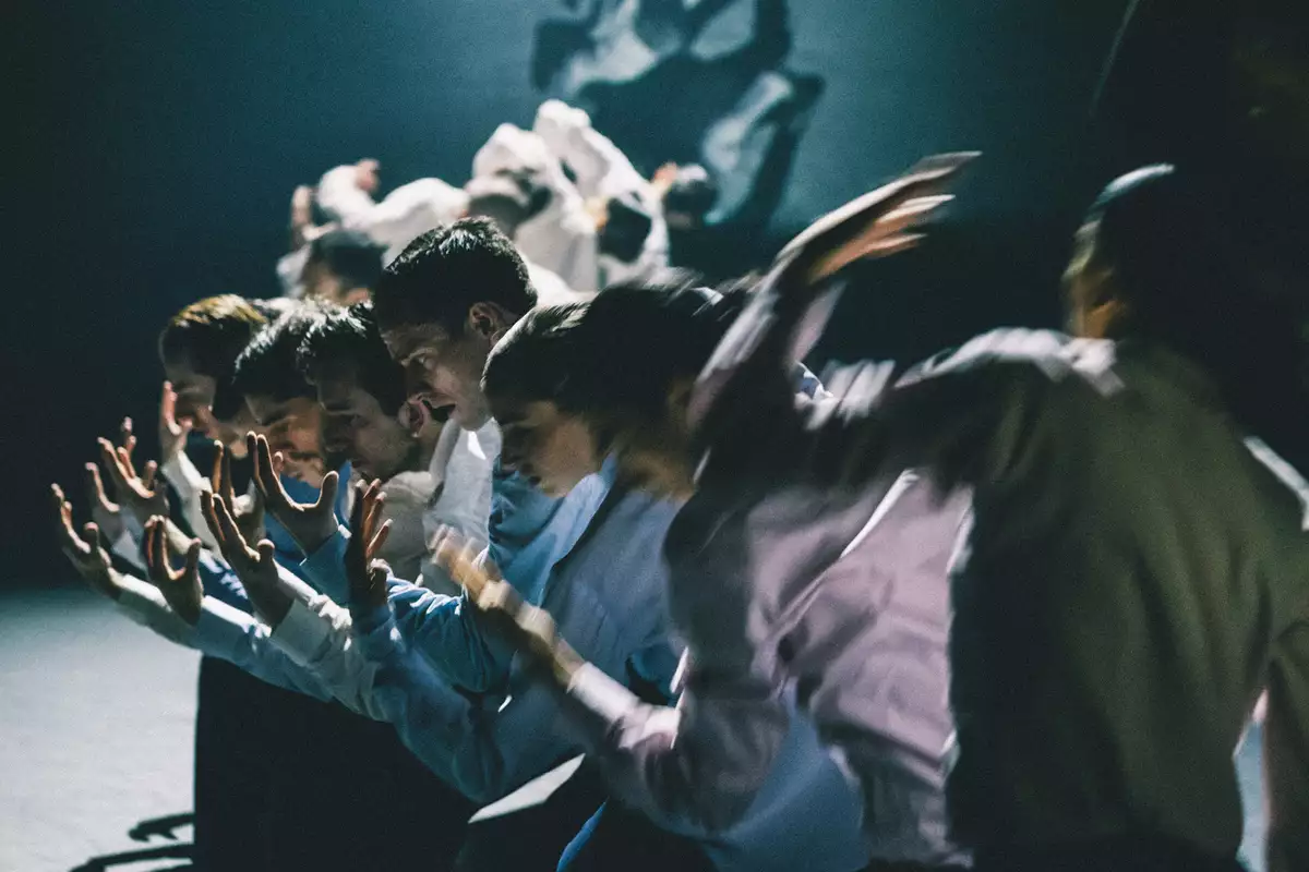 A group of dancers stood in a line with their arms extended forward, moving in blurred motion. The lighting is dim, casting dramatic shadows.