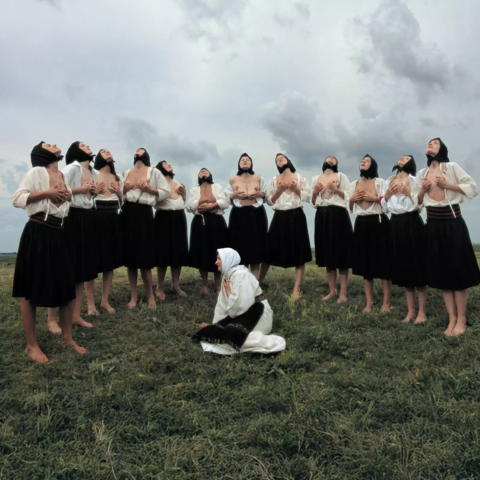 A group of women in traditional Eastern European attire stand barefoot in a grassy field under a dramatic, cloudy sky. Most are wearing black skirts, white blouses, and black headscarves, with some partially exposing their chests while lifting their heads upward in a ritualistic pose. In the foreground, a single woman dressed in white kneels on the ground with a headscarf and embroidered belt, gazing to the side.