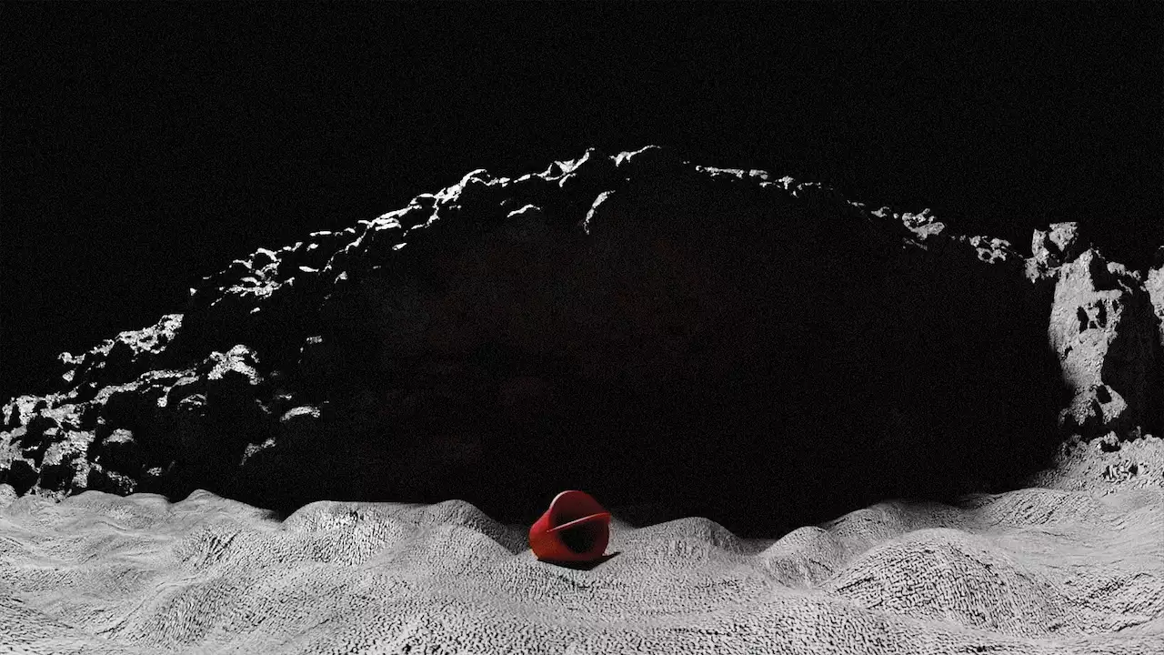 A black-and-white photograph of sand and rocks with a red bucket in the centre.