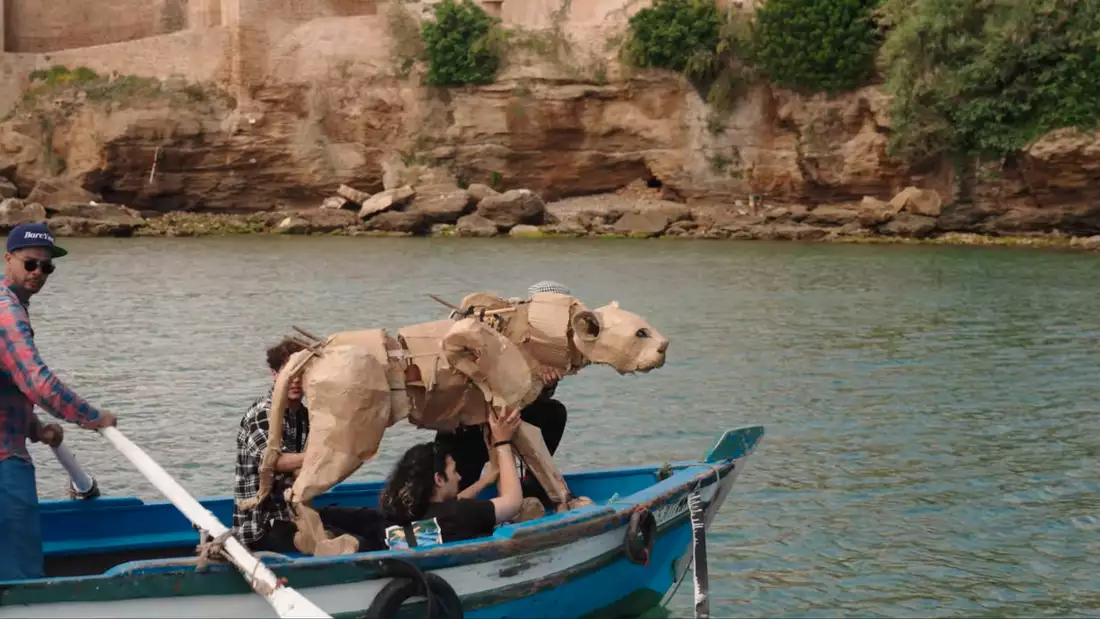 Puppet animals on boats with puppeteers in bright blue sea. There is a port behind them.