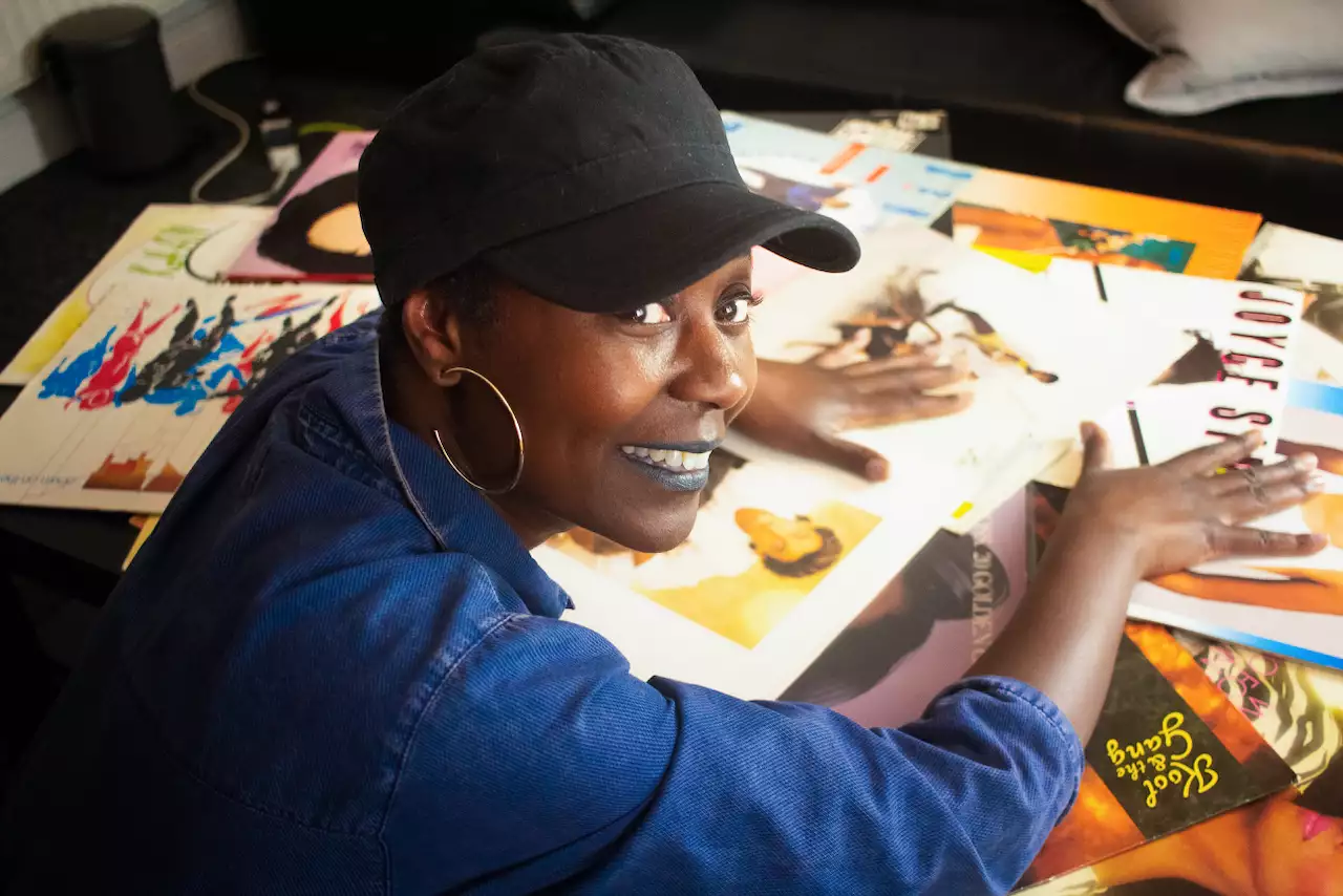 A person wearing hooped earrings and a black cap smiles at the camera. Their hands are placed on a table covered with records.