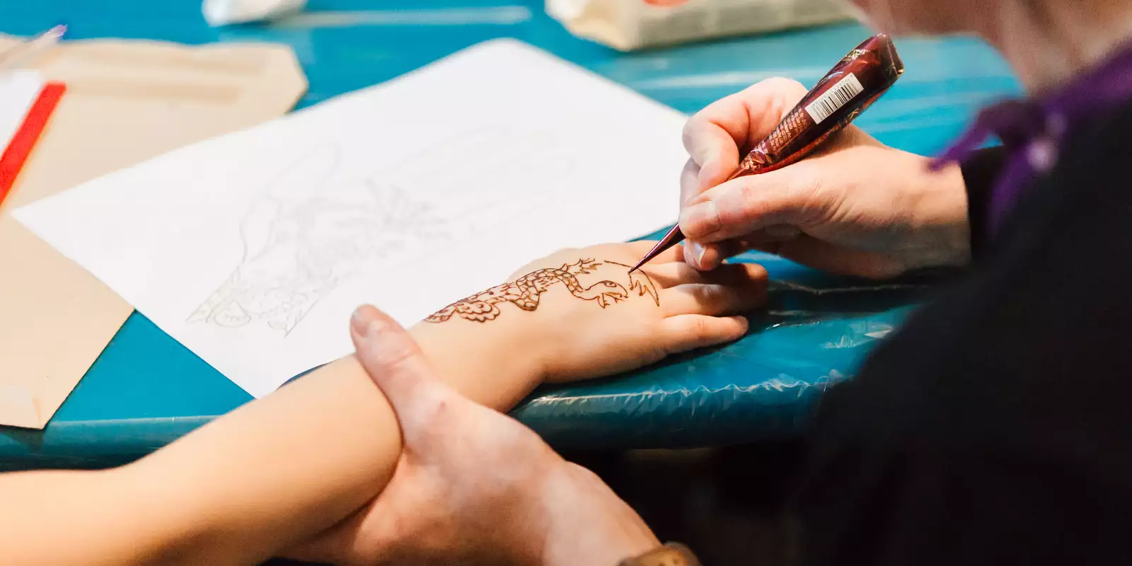 A person (out of frame) lays their right arm on a table while an artist draws a henna tattoo on the back of their hand