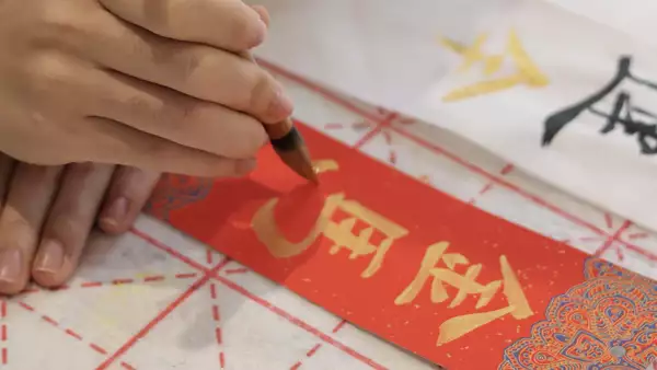 Hand writing Chinese calligraphy in gold ink on a red paper.