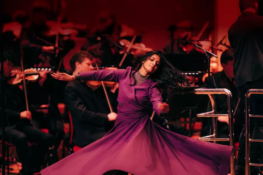 A dancer in a flowing purple dress gracefully spins on stage. Behind her, an orchestra dressed in black plays classical instruments.