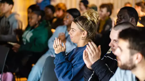 A room of people clapping. In the centre is a woman wearing a blue jumper clapping.
