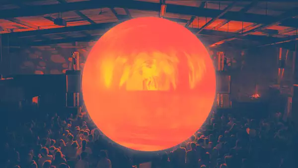 A large, glowing orange orb is suspended above a crowd in a dark indoor venue, casting warm light over the audience and the industrial-style ceiling.
