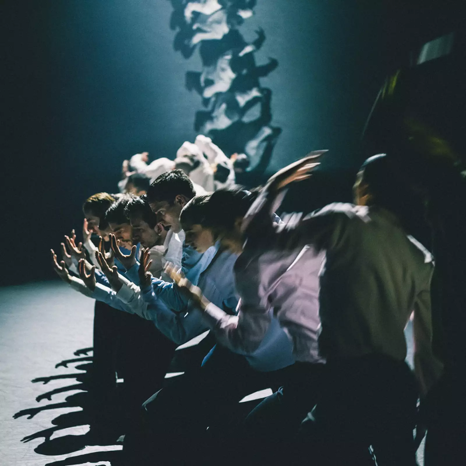 A group of dancers stood in a line with their arms extended forward, moving in blurred motion. The lighting is dim, casting dramatic shadows.