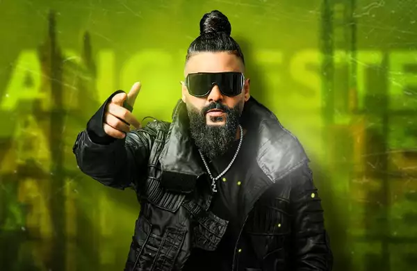 Sasy with his hair in a bun wearing dark sunglasses. The background is green with the word 'Manchester' displayed three times.