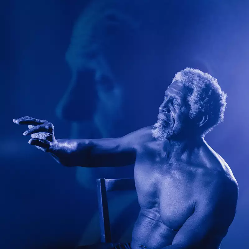 A shirtless elderly man with curly white hair and a beard sits on a chair, reaching out with an expressive hand. The image has a blue monochromatic tone with a soft-focus overlay of his face in the background.