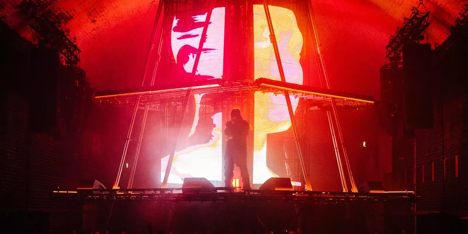 A photograph of Skepta, stood in silhouette on a brightly lit stage. The stage is a custom-built pyramid with a flat top, with a centrepiece screen showing 3D graphics of a human head shown across multiple large LED displays