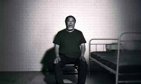 Black-and-white image of Ai Weiwei sitting on a chair beside a simple metal bed against a brick wall.