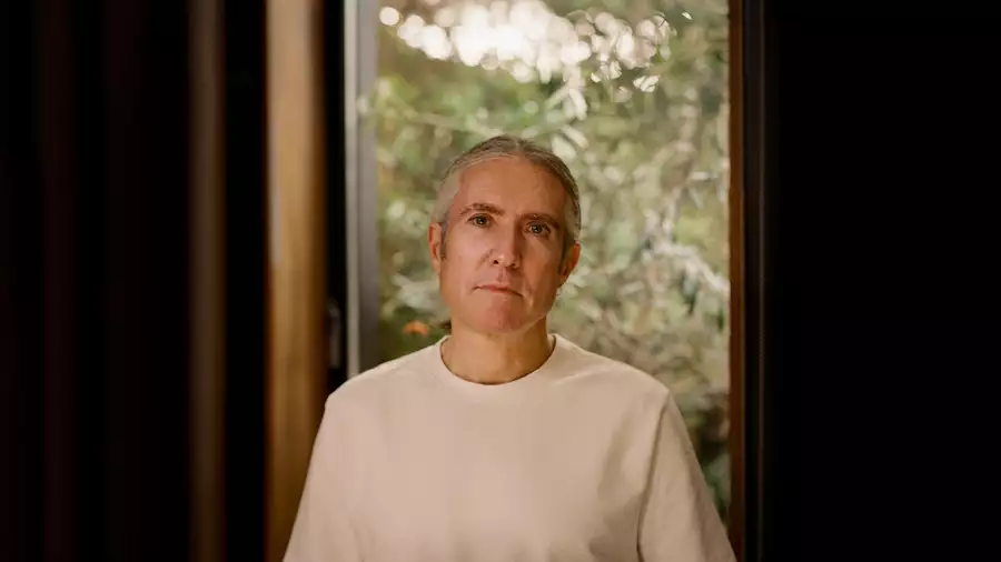 A portrait of Richard Russell wearing a light-coloured shirt, standing in front of a window with a blurred green outdoor background.