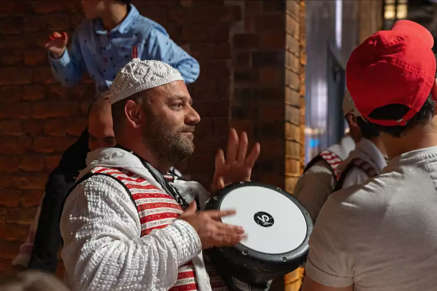 A man in traditional attire plays a hand drum at a lively cultural event, surrounded by people enjoying and dancing to the performance.