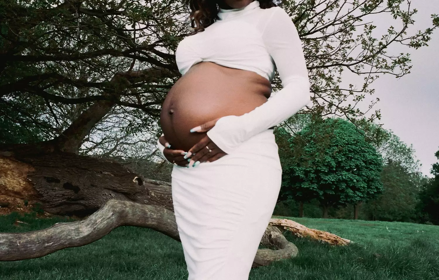 A pregnant woman standing in a green space with trees. She is wearing a white crop top and skirt, and she is cradling her stomach. Her head is not visible.