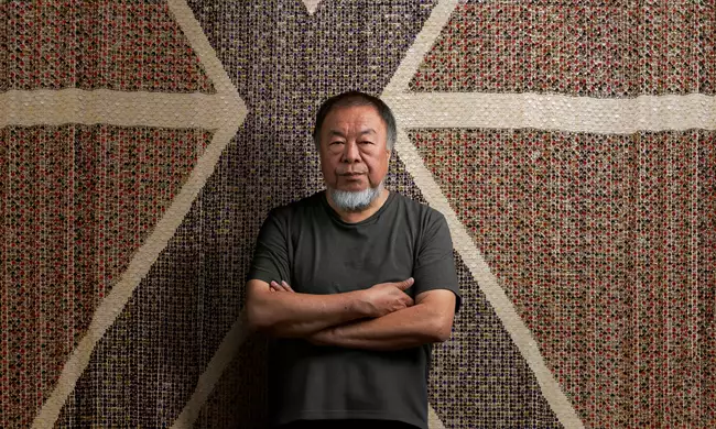 Ai Weiwei stood with arms folded wearing a black t-shirt, in front of a giant flag made of buttons