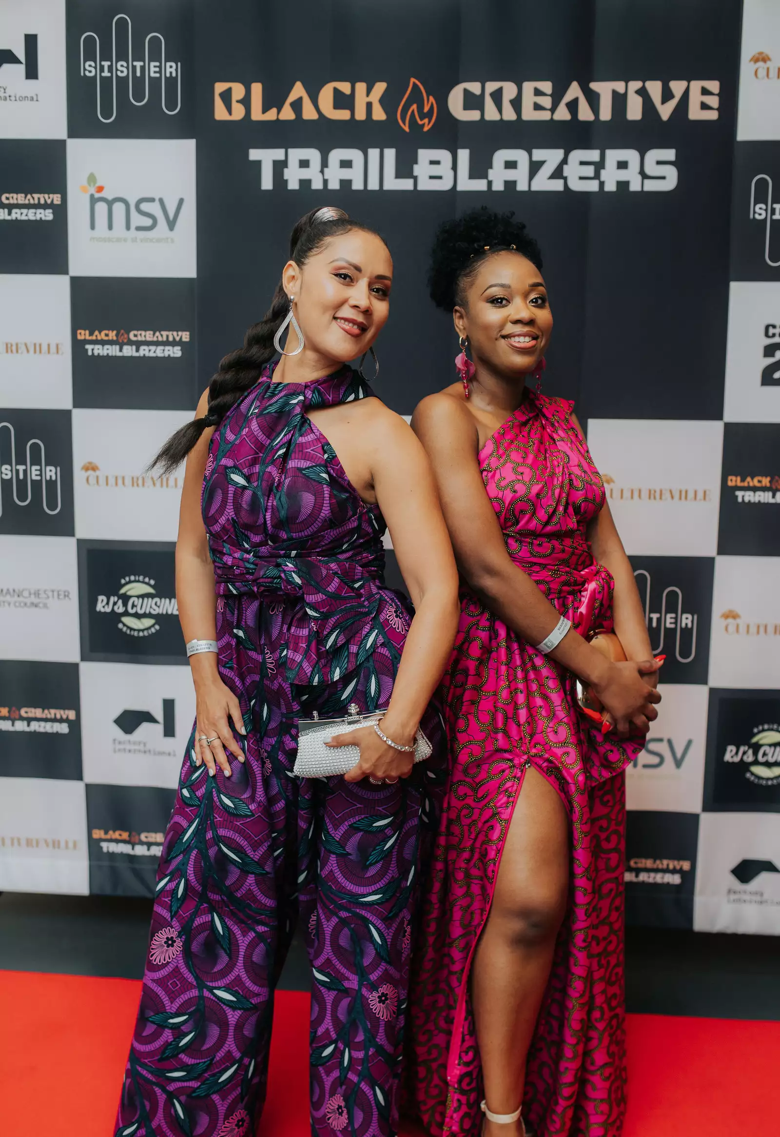 Two women pose confidently on the red carpet at the "Black Creative Trailblazers" event, wearing vibrant African-inspired outfits in shades of purple and pink. Behind them is a branded backdrop.